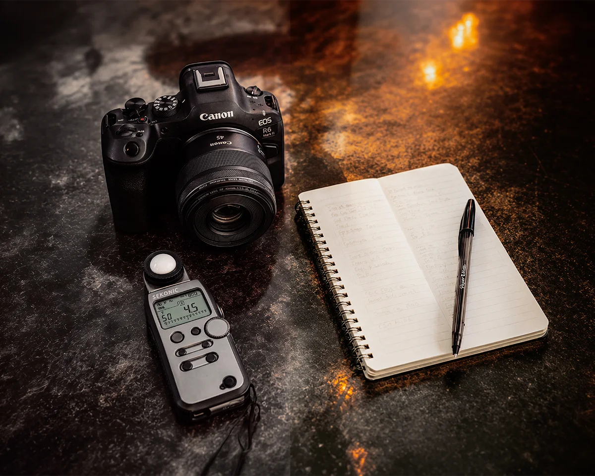Canon camera, light meter, notebook, and pen arranged on a dark tabletop.