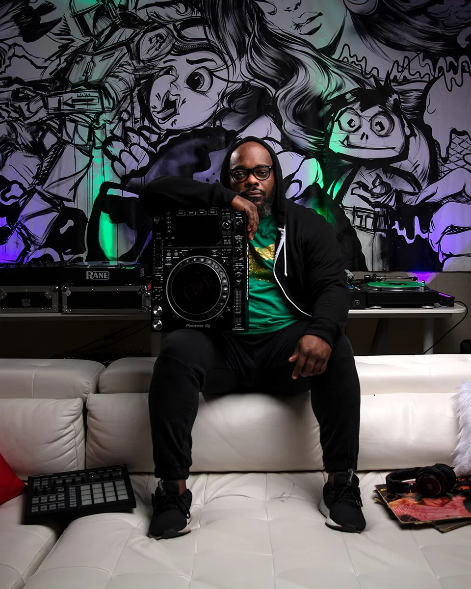 DJ Tillery James seated with a cinema camera in a mural-covered studio during a promotional shoot.